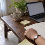 Hands write on a notepad beside an open laptop, a mug of tea, sticky notes, and shoes on a tiled floor by a window.